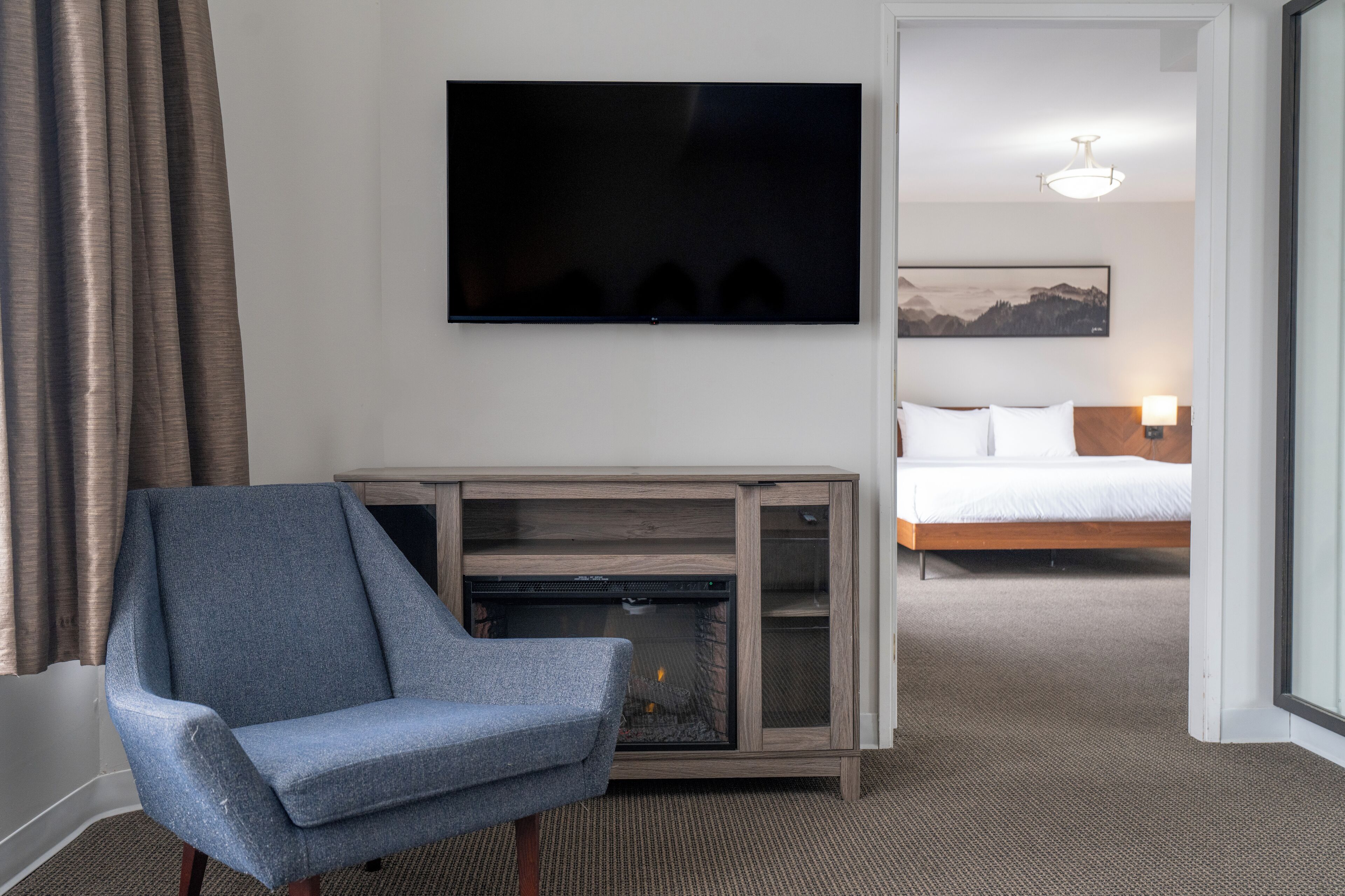 A hotel room with a flat-screen TV mounted on the wall, a seating area with a blue armchair, a fireplace, a bed with white bedding, and a framed picture on the wall.
