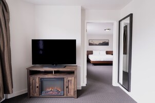 Habitación de hotel con televisión sobre un soporte de madera, chimenea, cama con ropa de cama blanca y un cuadro en la pared.