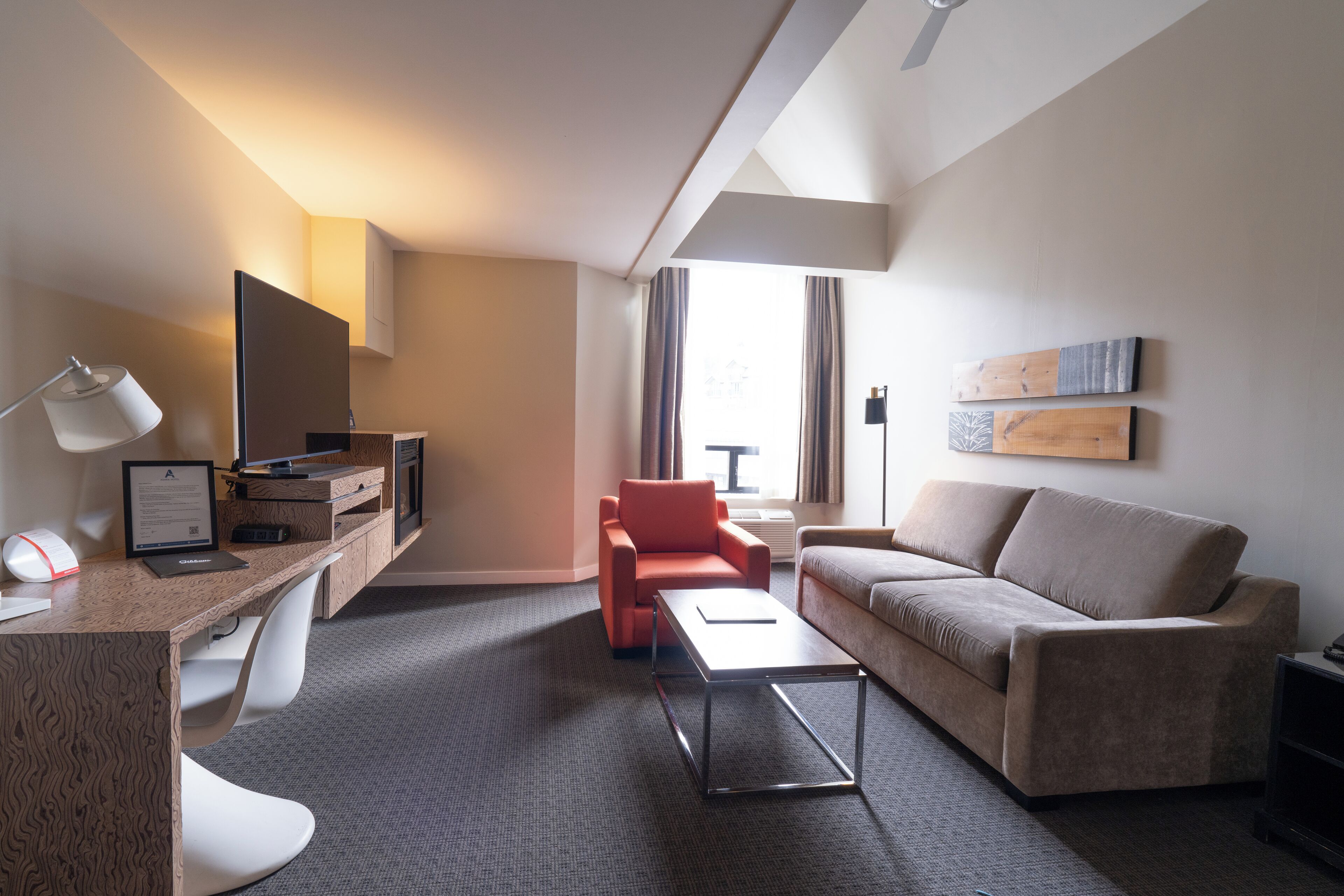A modern hotel room with a desk, a red armchair, a sofa, and a flat-screen TV.