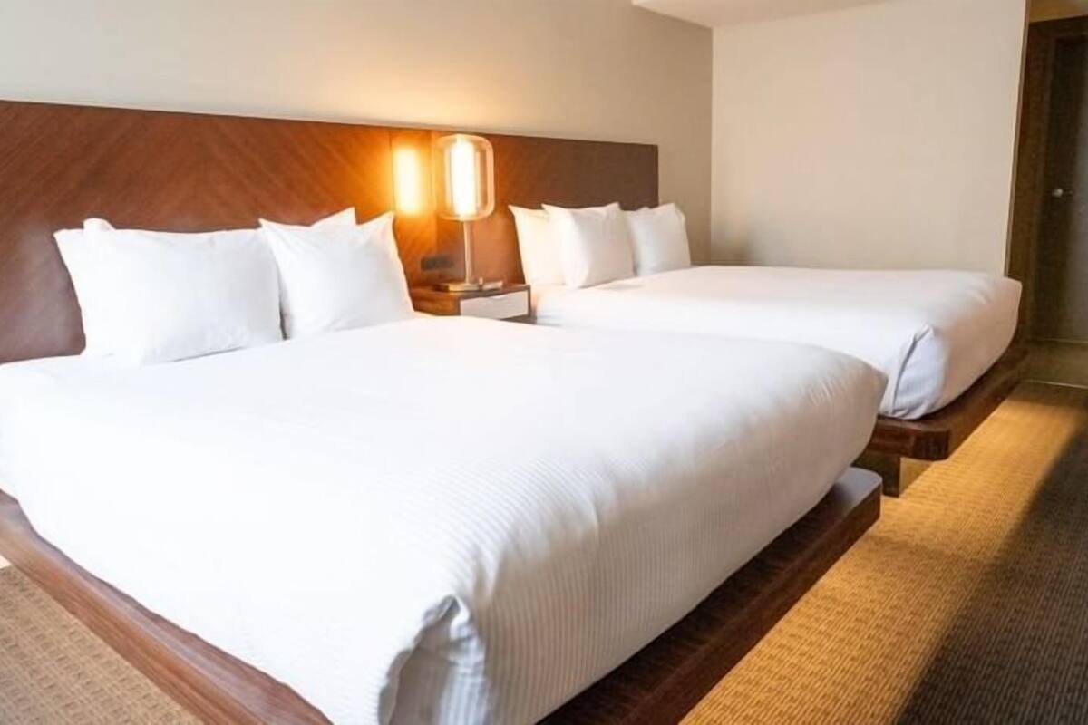 A hotel room with two beds, a wooden headboard, a nightstand with a lamp, and a carpeted floor.
