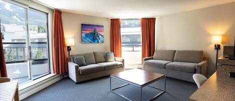 A hotel room with two sofas, a coffee table, and a TV. There are windows with curtains, a painting on the wall, and a view of buildings outside.