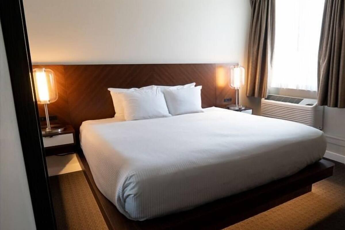 A hotel room with a large bed, two bedside tables with lamps, a wall-mounted air conditioner, and a window with curtains.