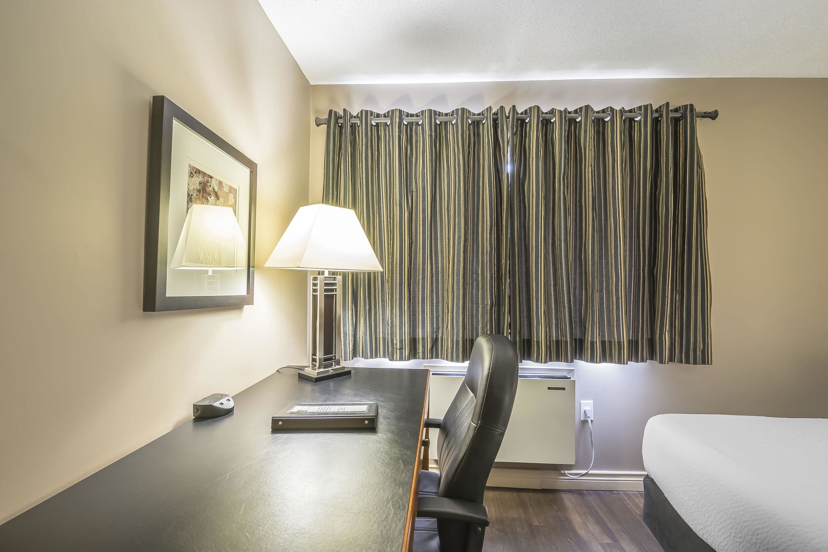Habitación de hotel con una cama, un escritorio con lámpara, una silla, un pequeño escritorio y un ventanal con cortinas.