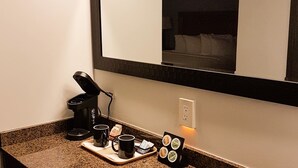 A hotel room with a coffee machine, two mugs, a tray with coffee pods, and a bed visible in the mirror reflection.
