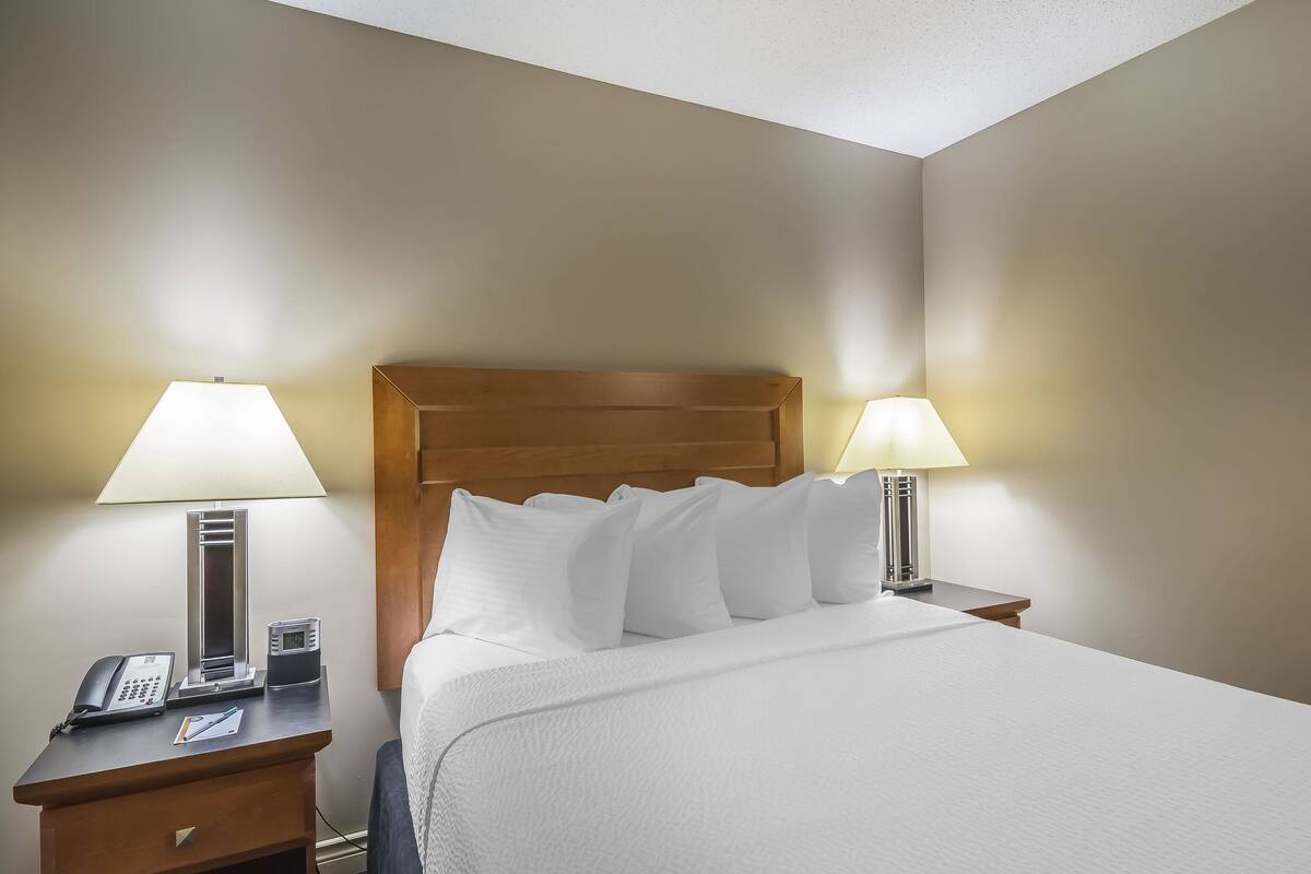 A hotel room with a wooden headboard, white bedding, two bedside lamps, a bedside phone, and a small table with a lamp.