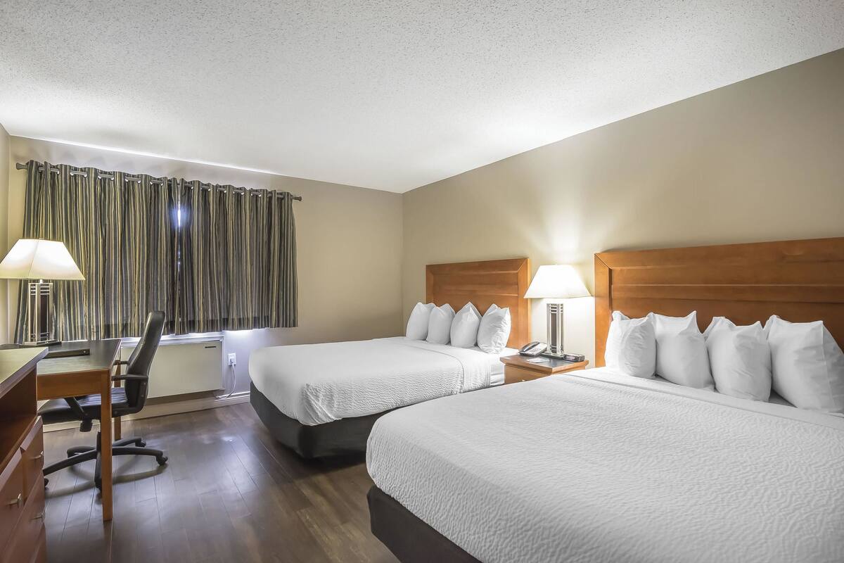 A hotel room with two beds, a desk with a chair, a lamp, and a window with curtains.
