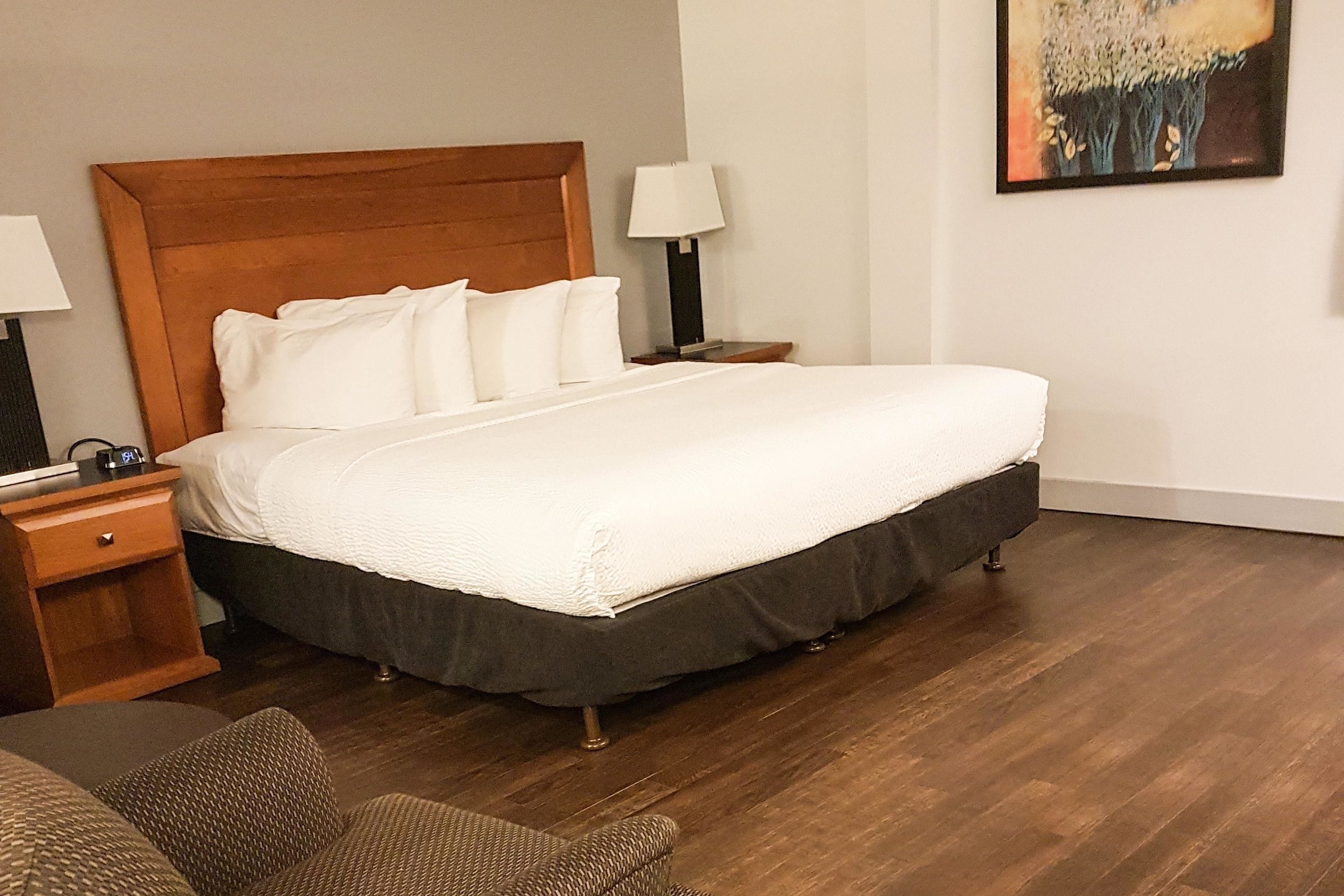 Una cama bien hecha con tablero de madera, una mesita de noche con lámpara y una silla en una habitación con suelo de madera.