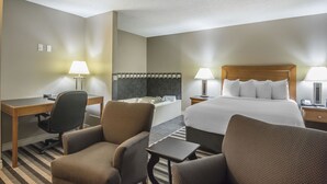 A hotel room with a bed, two chairs, a desk, a lamp, a small table, and a bathtub.