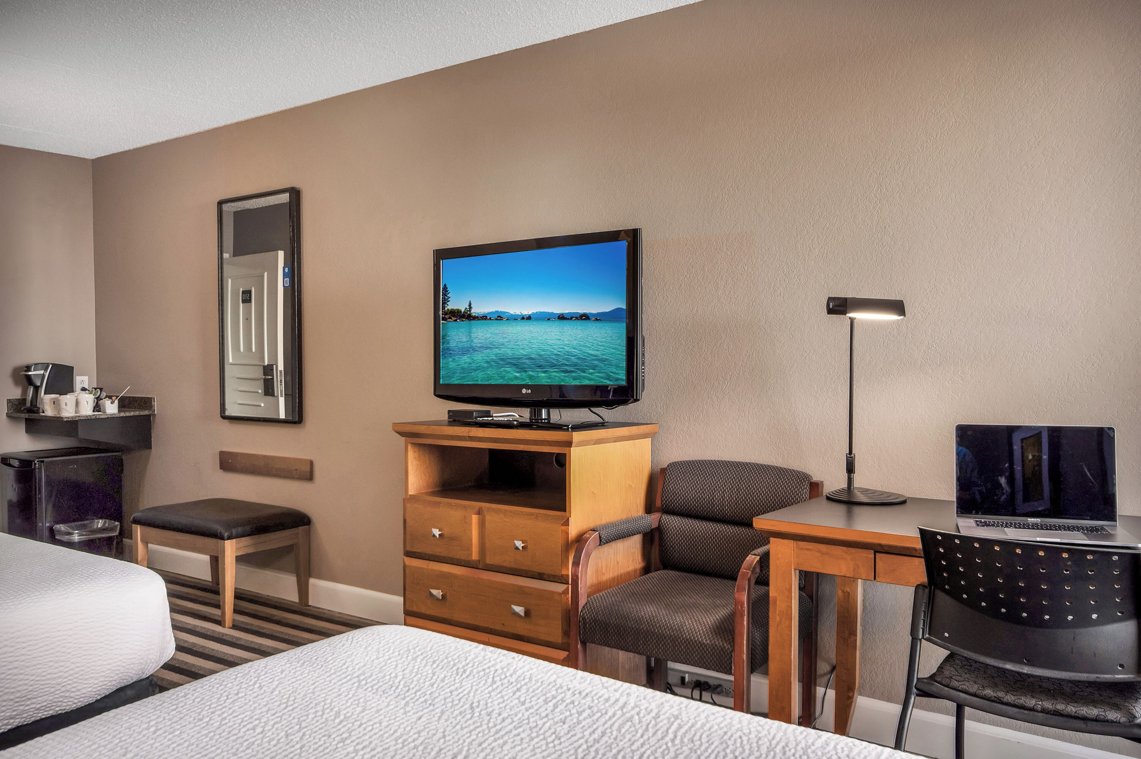 A hotel room with a bed, a desk with a laptop, a television, a chair, a desk lamp, and a small table with a coffee maker.