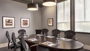 A conference room with a round table, six chairs, framed pictures on the wall, and large windows with blinds. - Royal Hotel Regina, Trademark Collection by Wyndham (Regina)