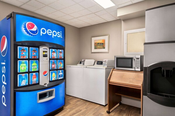 Una máquina expendedora con la marca Pepsi, un microondas y un refrigerador en una habitación con suelo de madera y un cuadro en la pared.