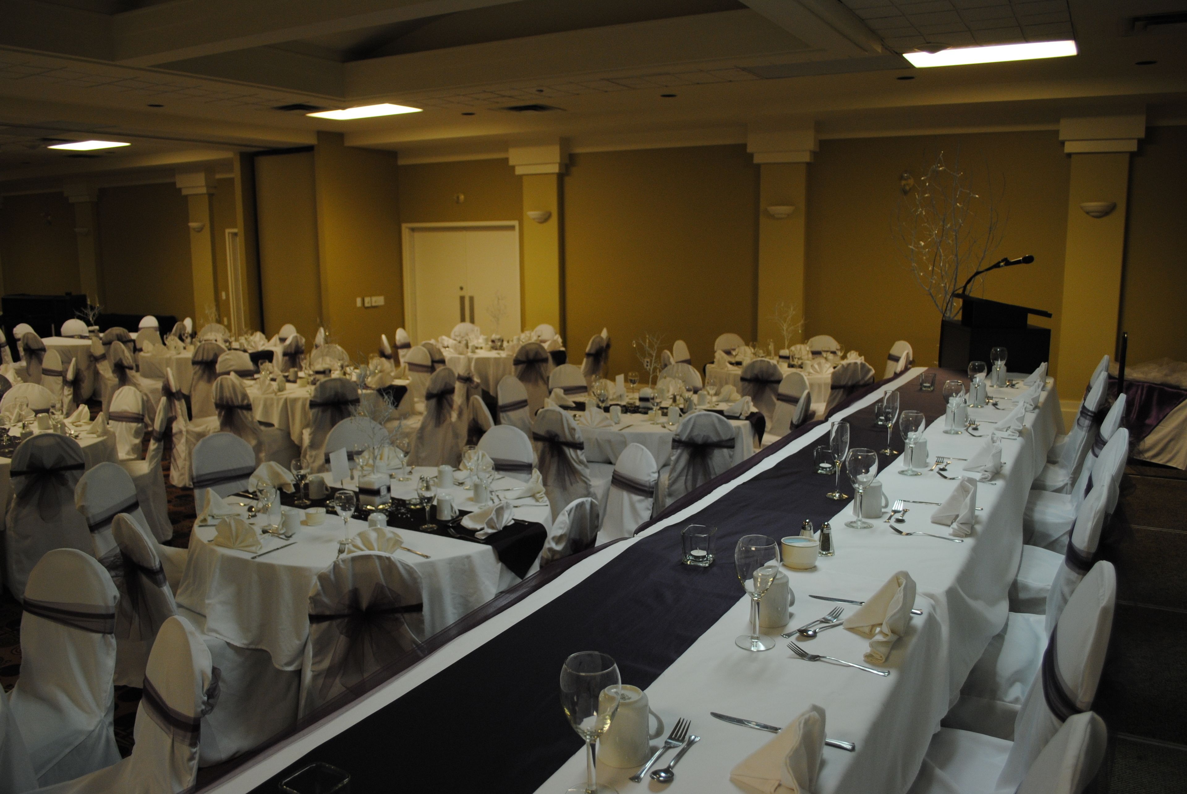 Une salle de banquet où de longues rangées de tables sont dressées pour un événement formel, chacune étant recouverte d’une nappe blanche et agrémentée de chaises.