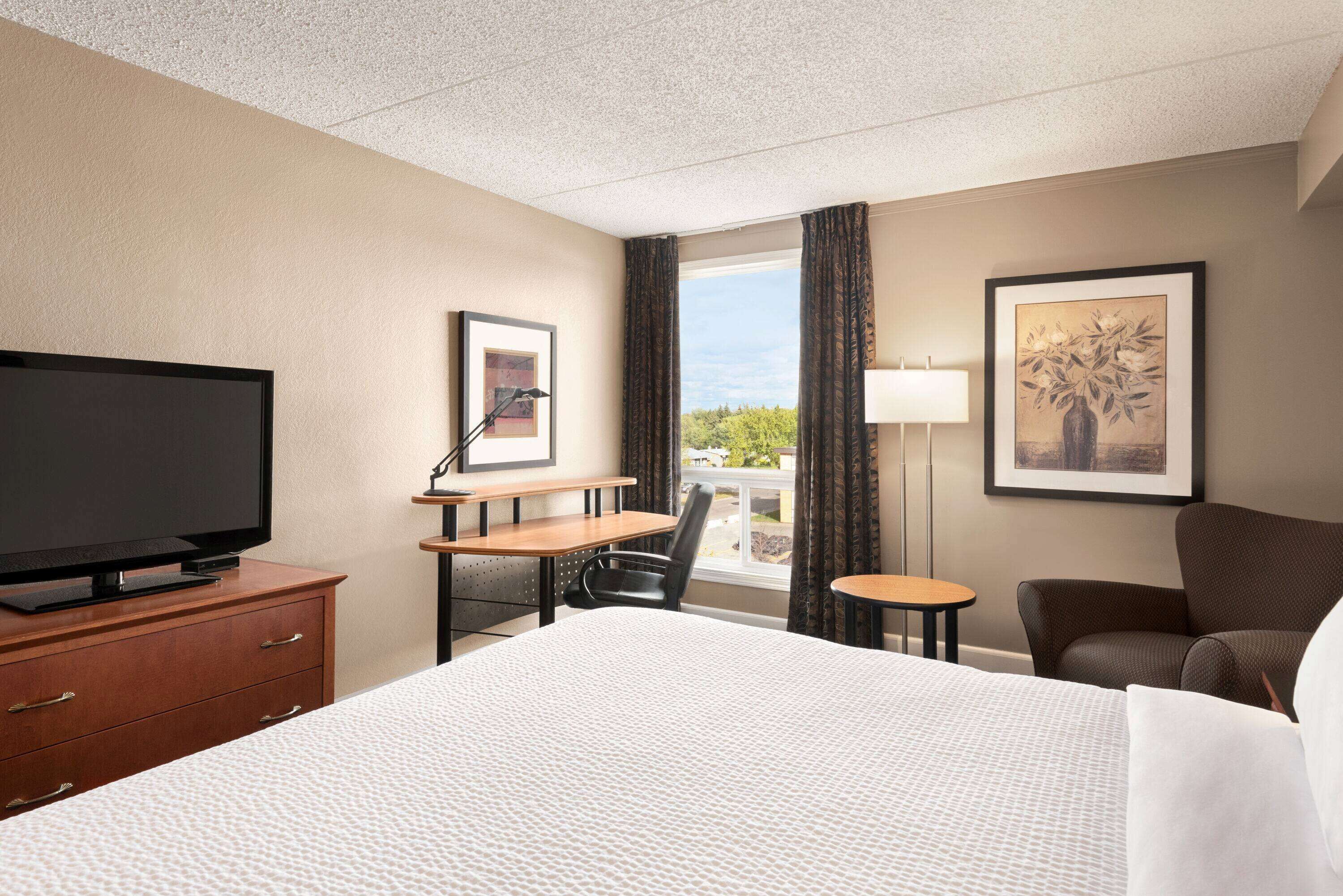 Habitación de hotel con una cama grande, un escritorio con silla, televisión, ventana con cortinas y obras de arte en las paredes.