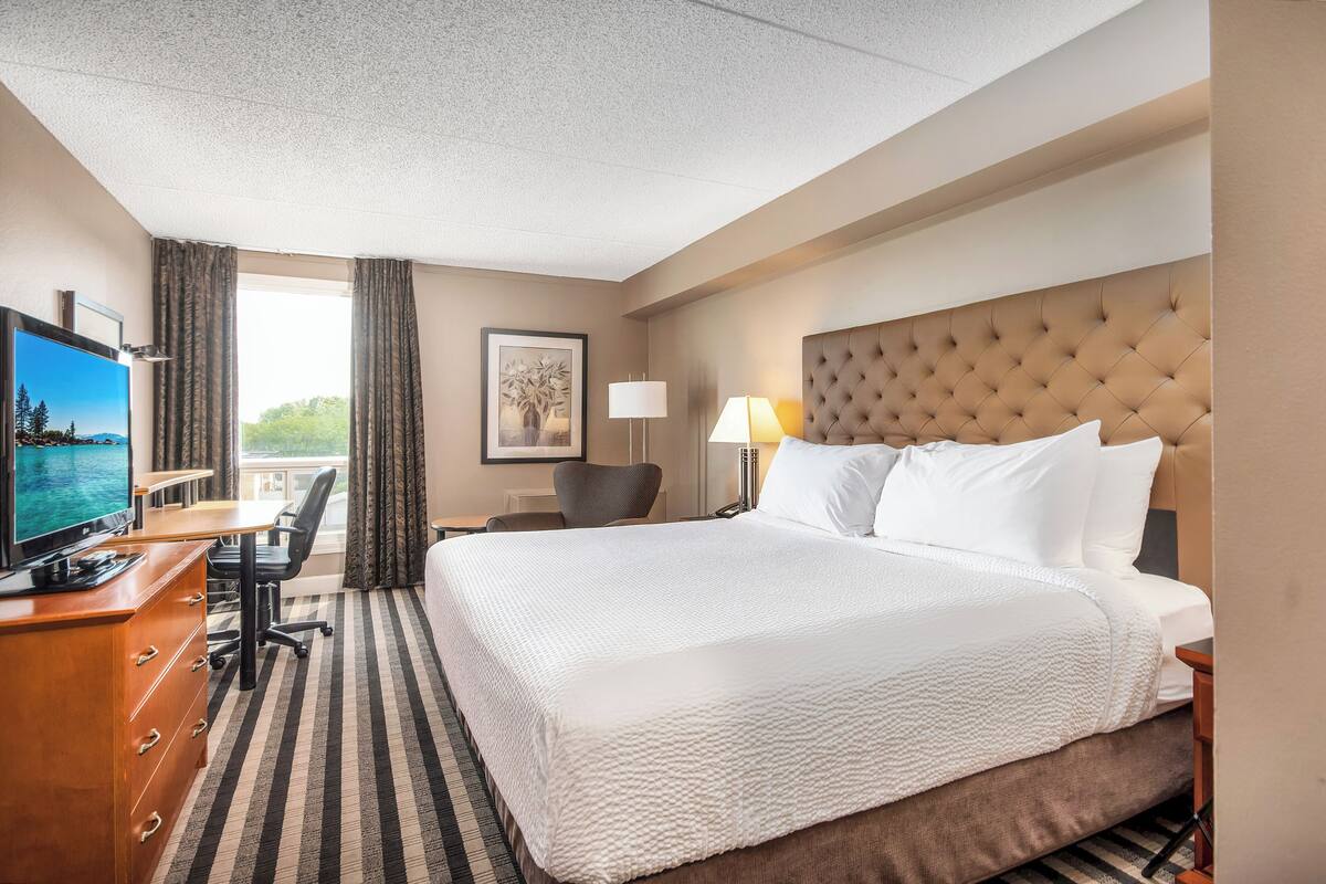 Ein Hotelzimmer mit einem großen Bett, einem Schreibtisch mit Fernseher, einem Sessel, einer Lampe und einem Fenster mit Vorhängen.