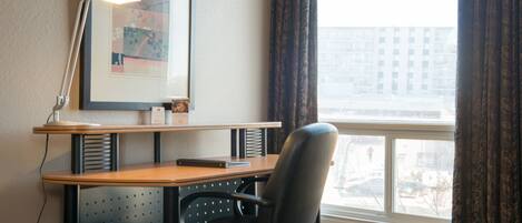 Une chambre d’hôtel équipée d’un bureau, d’une chaise, d’une lampe, d’un tableau encadré et d’une fenêtre avec des rideaux.