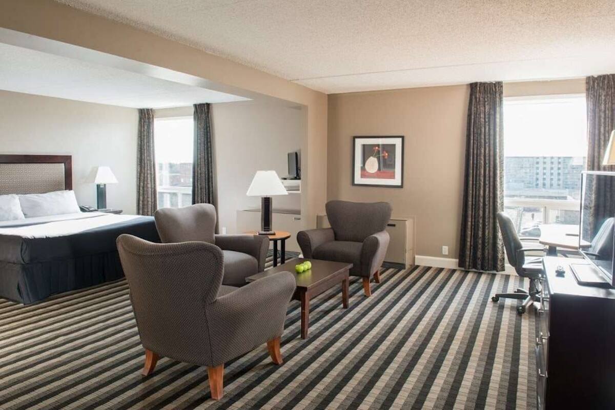 Ein Hotelzimmer mit einem Bett, zwei Sesseln, einem Schreibtisch, einer Lampe und Blick auf die Stadt.