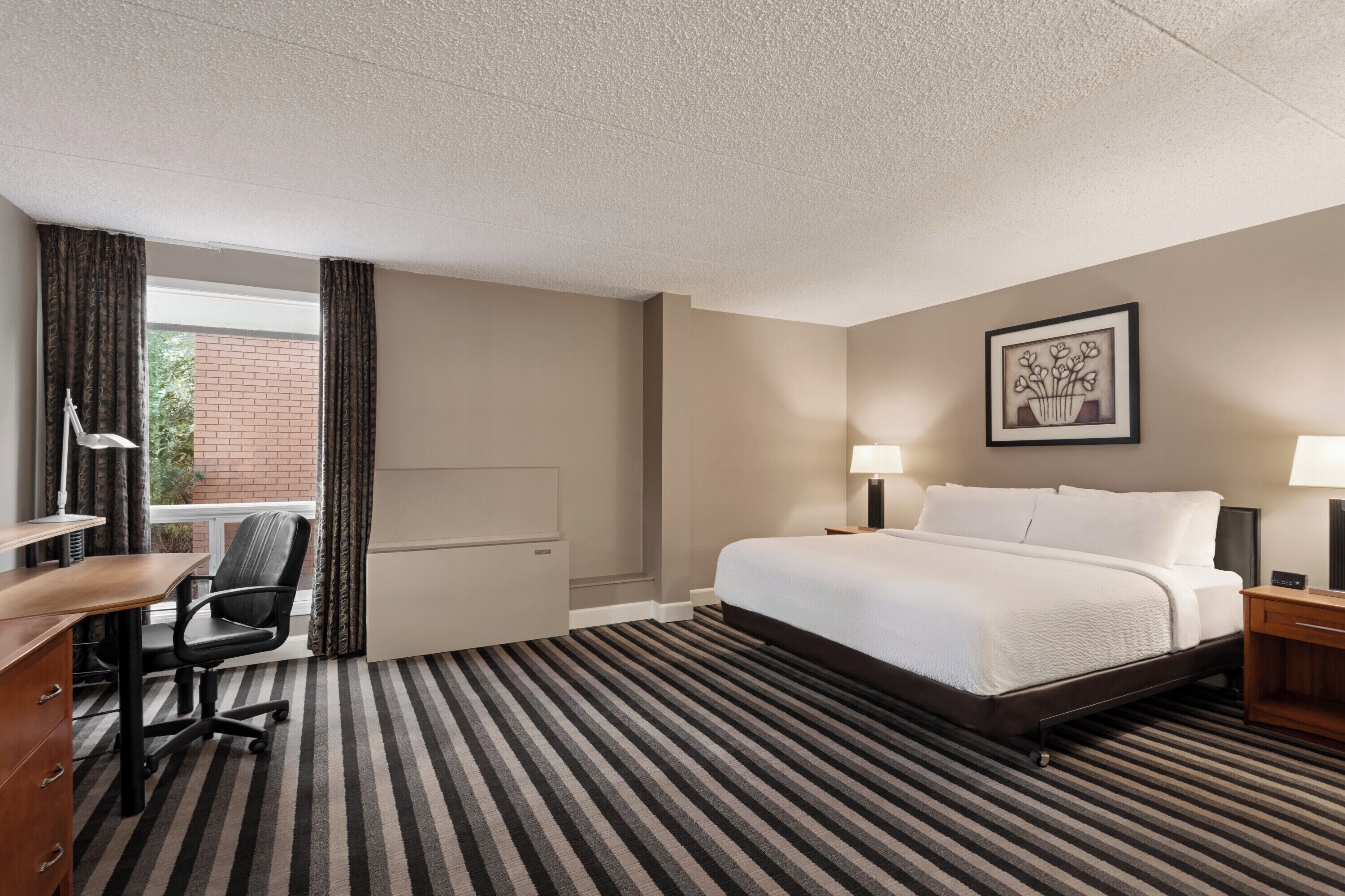 A hotel room with a bed, a desk, a chair, a lamp, and a framed picture on the wall.
