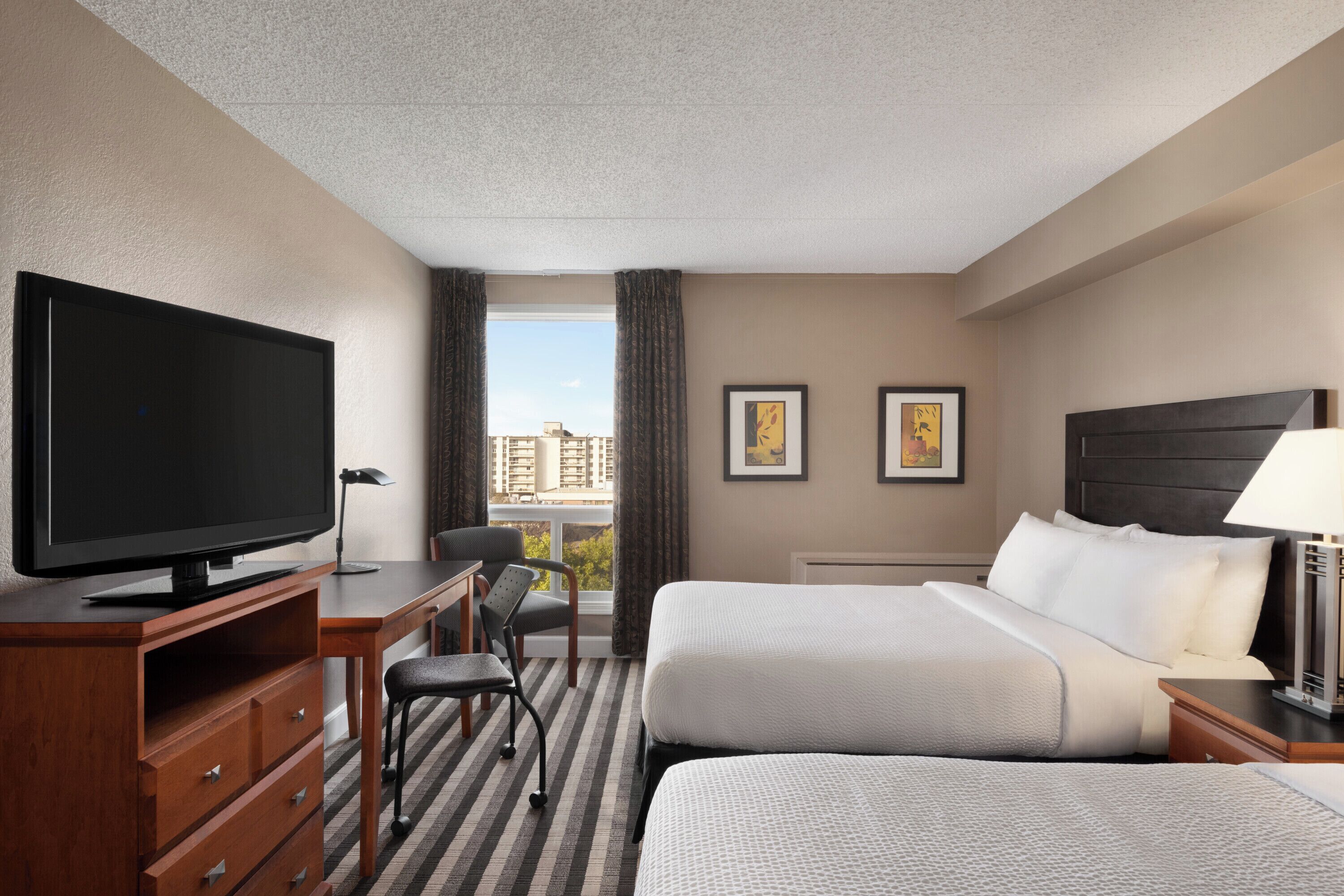 A hotel room with a television, two beds, a desk, a chair, and a window with curtains.