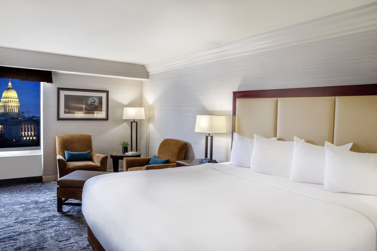 Habitación de hotel con una cama grande, dos sillones, una mesita de noche y vistas a un paisaje urbano.