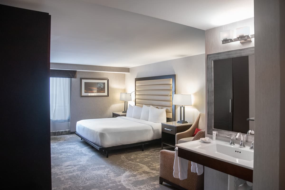 Una habitación de hotel con cama, mesita de noche, silla, espejo grande y baño con lavabo y espejo.