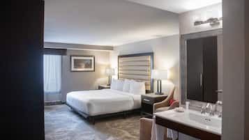 Una habitación de hotel con cama, mesita de noche, silla, espejo grande y baño con lavabo y espejo.