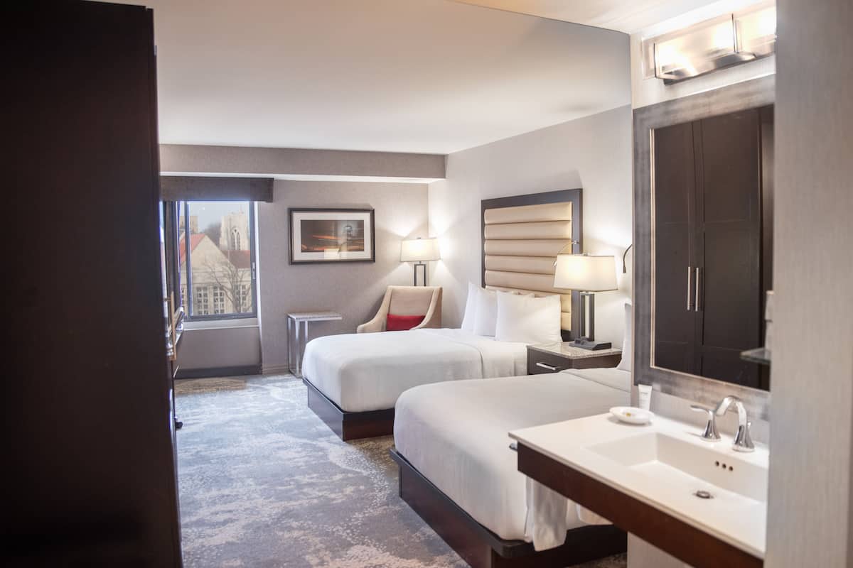 Habitación de hotel con dos camas, un espejo grande, un baño con lavamanos y vistas a un edificio a través de la ventana.