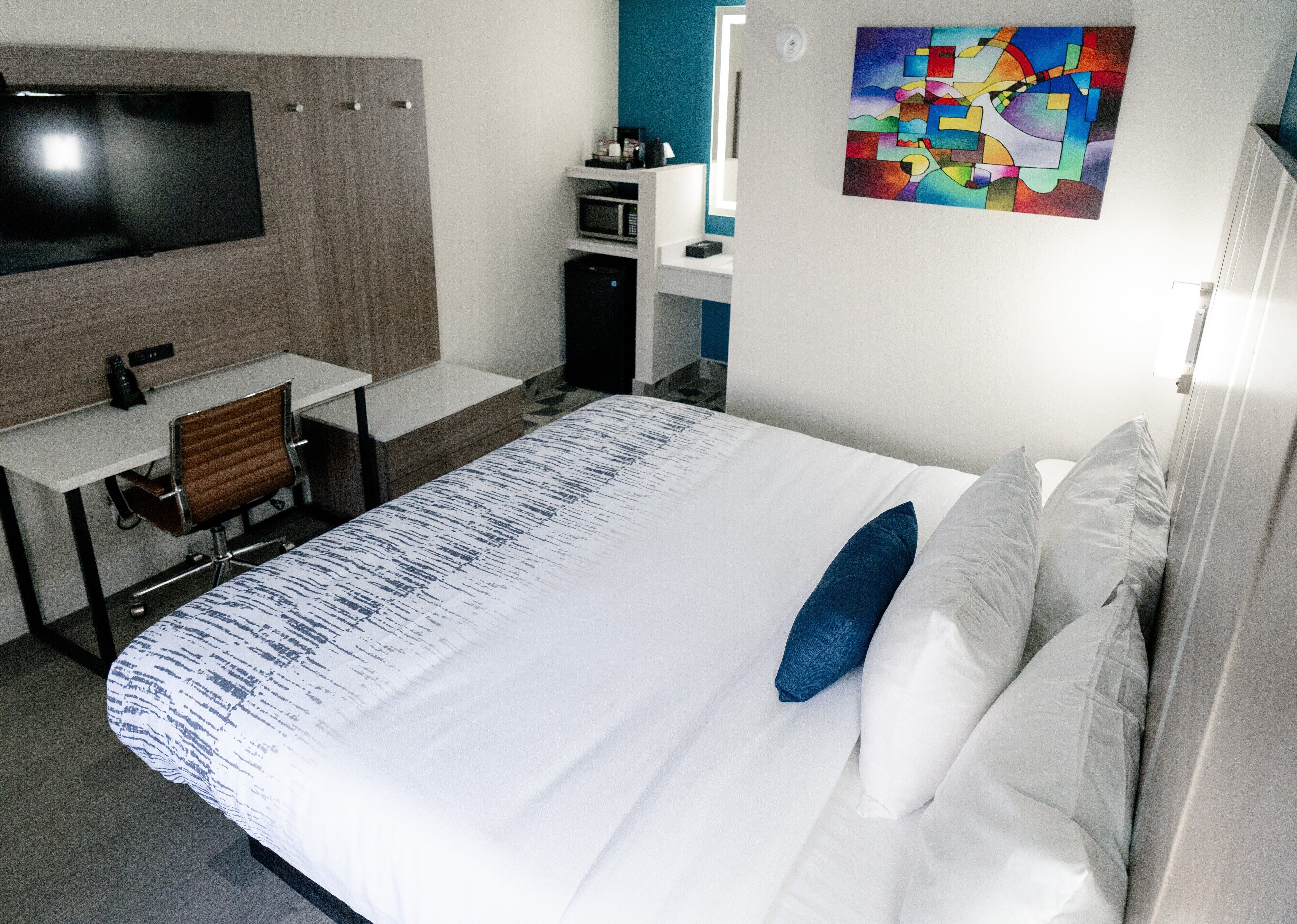 A hotel room with a bed, a desk, a chair, a TV, a microwave, and a painting on the wall.