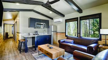 Une cuisine moderne avec des meubles sombres, un réfrigérateur en acier inoxydable et un four à micro-ondes. On y trouve une table à manger en bois avec des tabourets, un canapé en cuir et une grande fenêtre donnant sur les arbres.