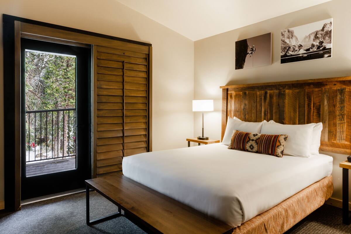 Habitación de hotel con una cama grande, cabecera de madera, un banco, una mesita de noche con lámpara y vista a árboles por una puerta corrediza.