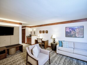 A hotel room with a flat-screen TV, a sofa, a chair, a desk with a lamp, and a painting of a canyon.