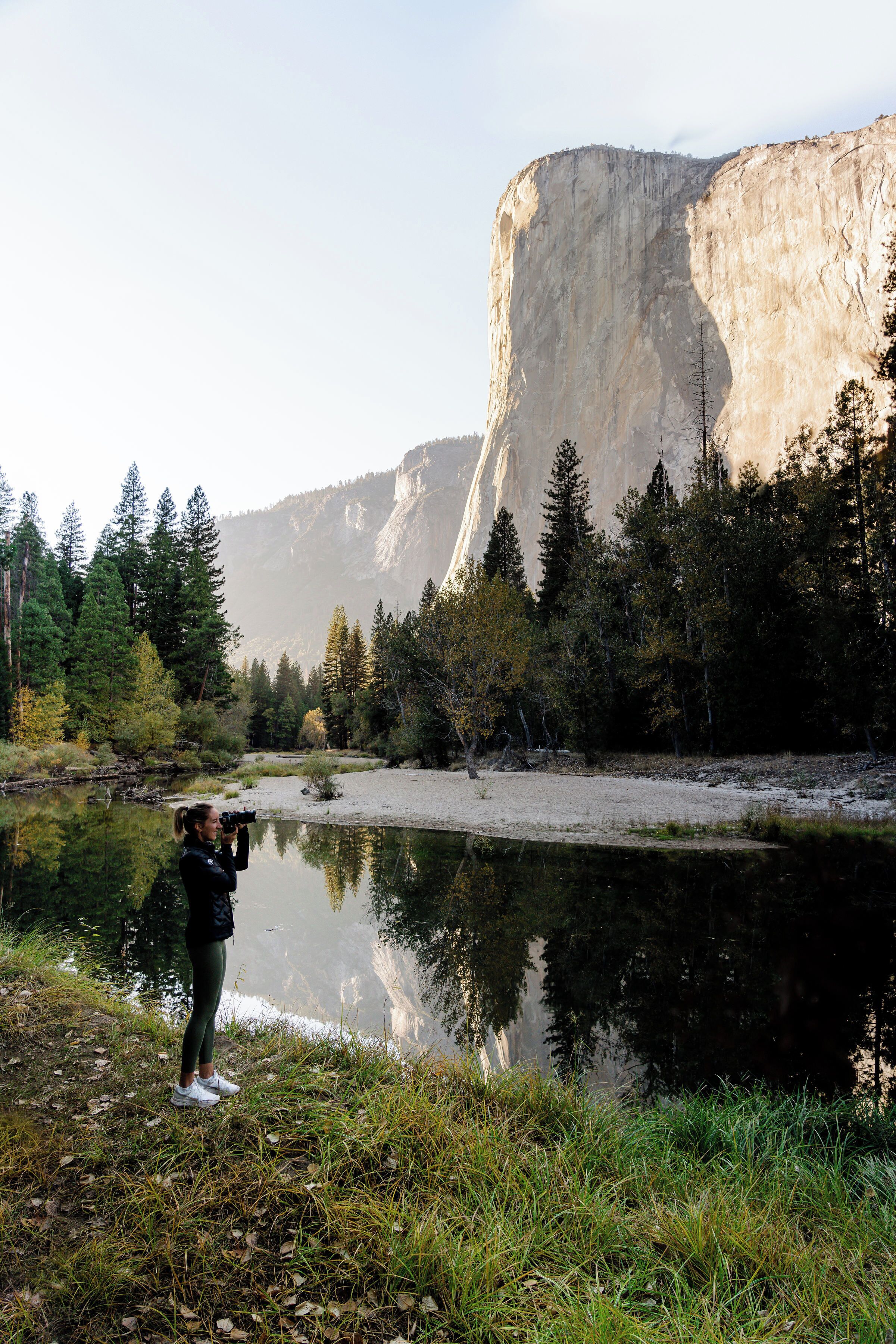 A person taking a photo near a calm river with a large cliff in the background.