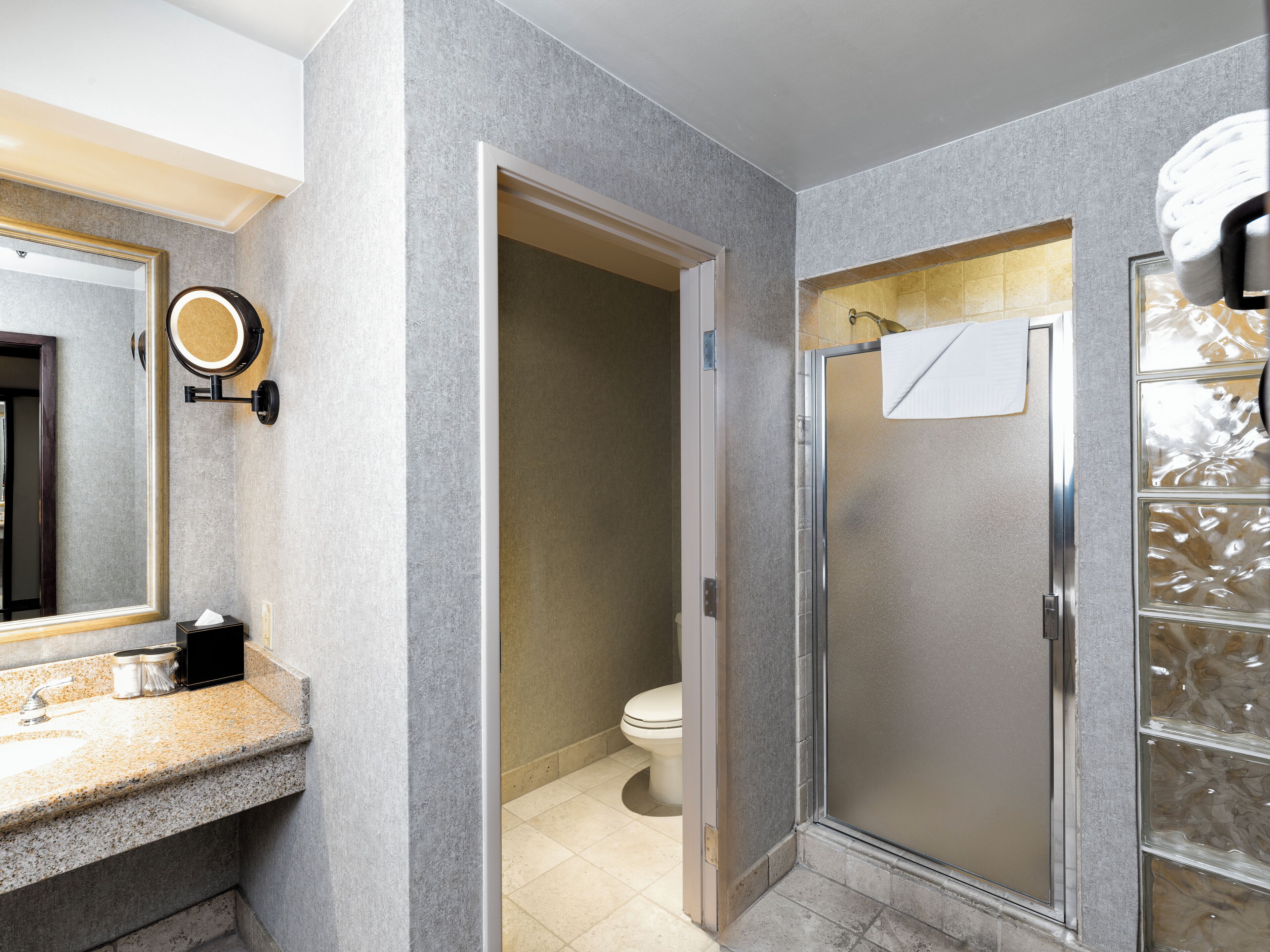 A modern bathroom with a granite countertop, a round mirror, a toilet, and a glass-enclosed shower.