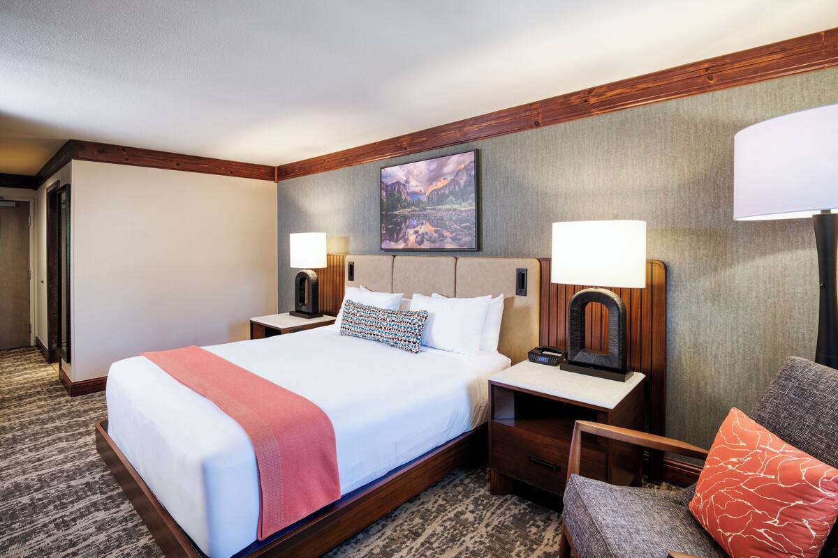 Una habitación de hotel con una cama grande, dos mesitas de noche con lámparas, una silla y un cuadro en la pared.