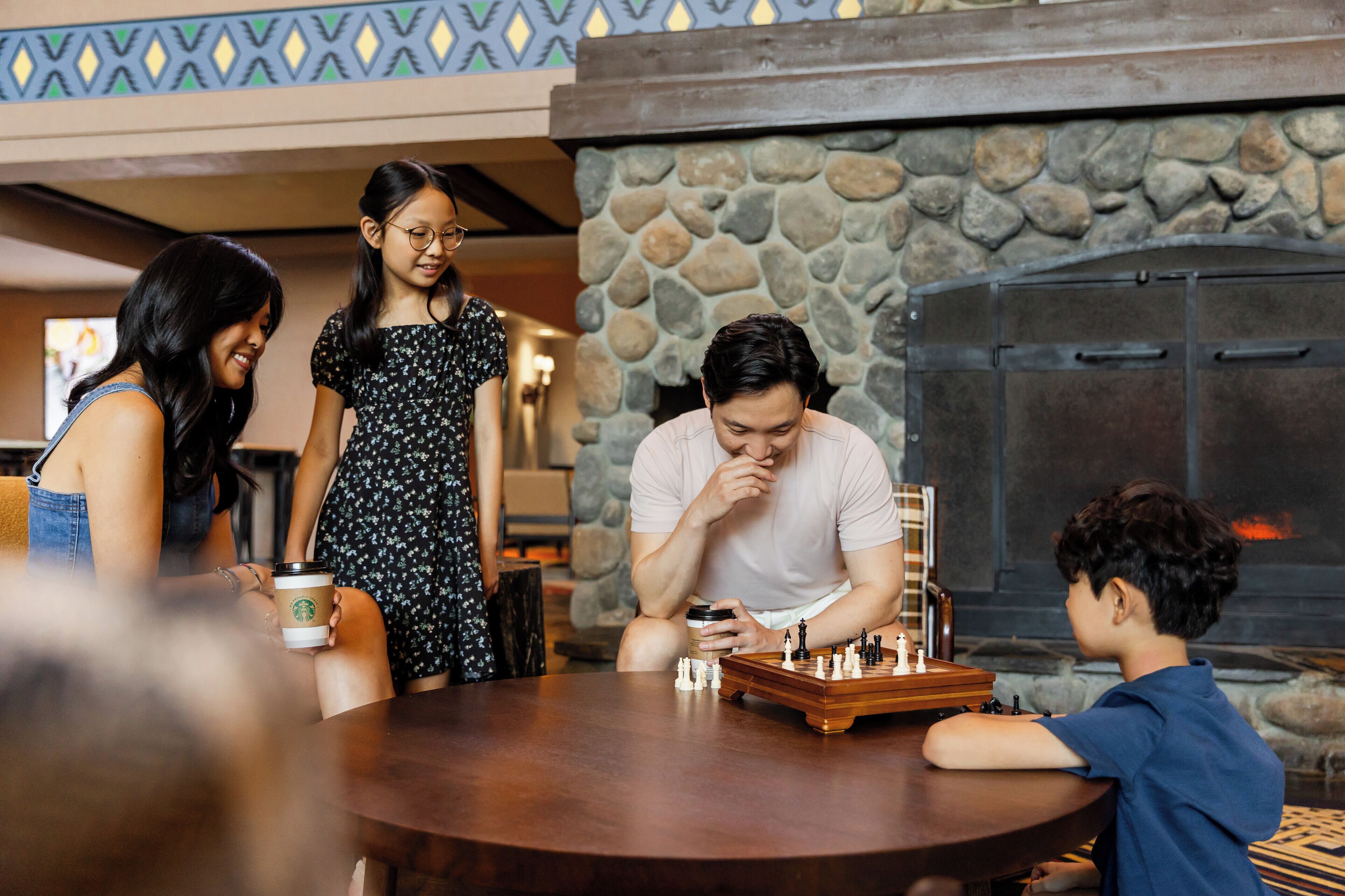 A group of people around a wooden table with a chessboard, in a room with a stone wall and a fireplace.