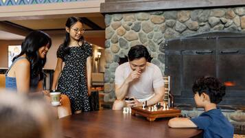 A group of people around a wooden table with a chessboard, in a room with a stone wall and a fireplace.