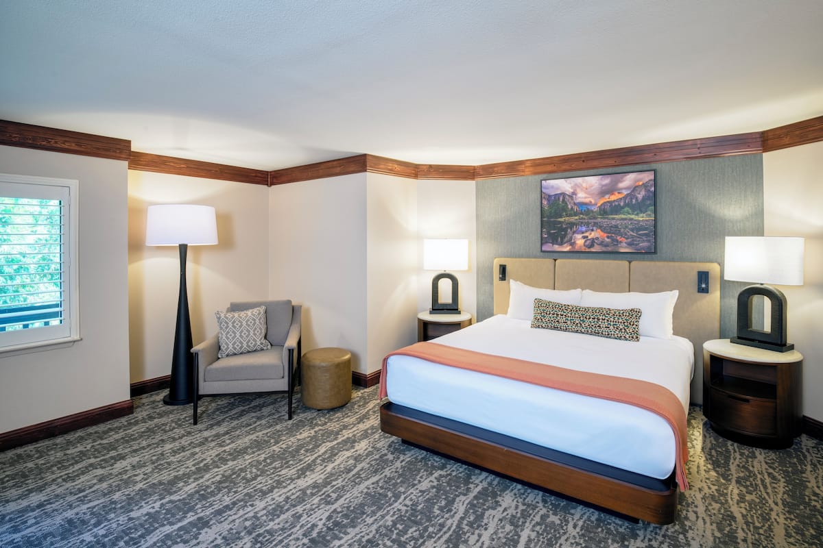 Habitación de hotel con una cama grande, dos mesitas de noche, una silla, una lámpara, un cuadro en la pared y una ventana con persianas.