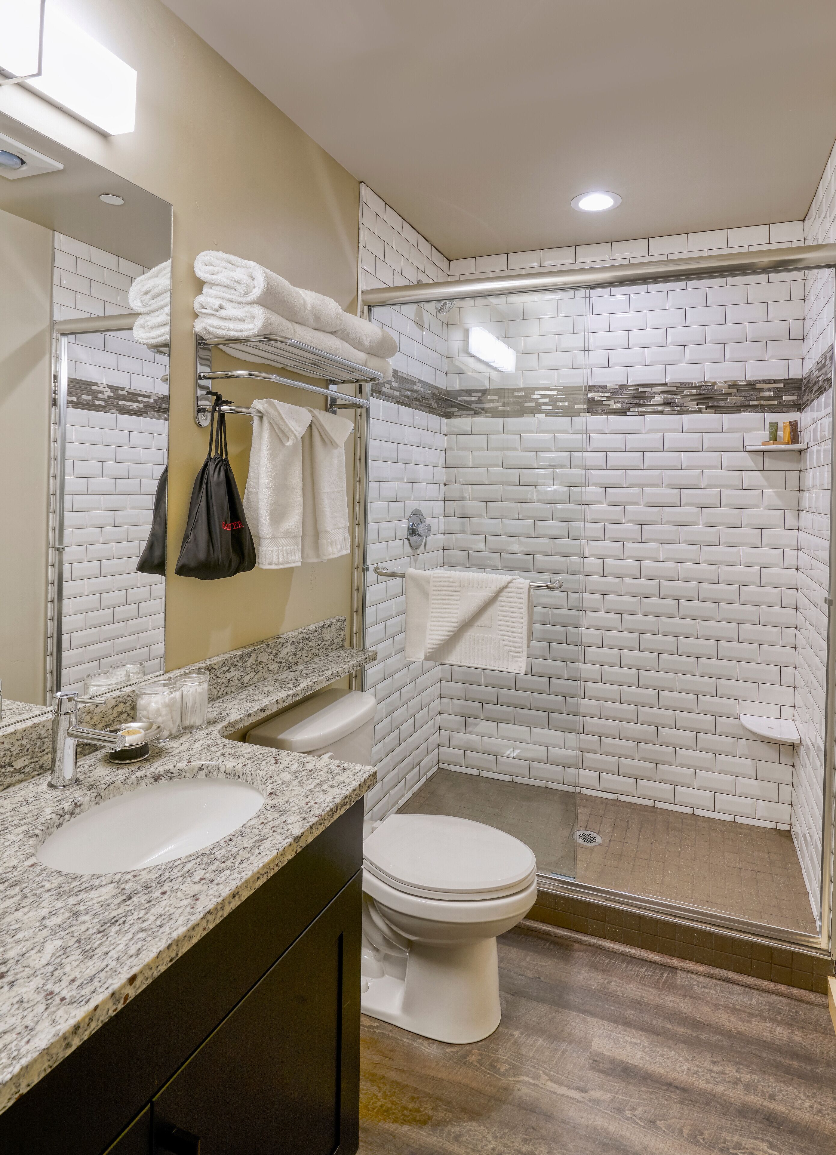 Een moderne badkamer met een granieten aanrechtblad, een wit toilet, een douche met glazen wand en een spiegel met handdoekhouder.