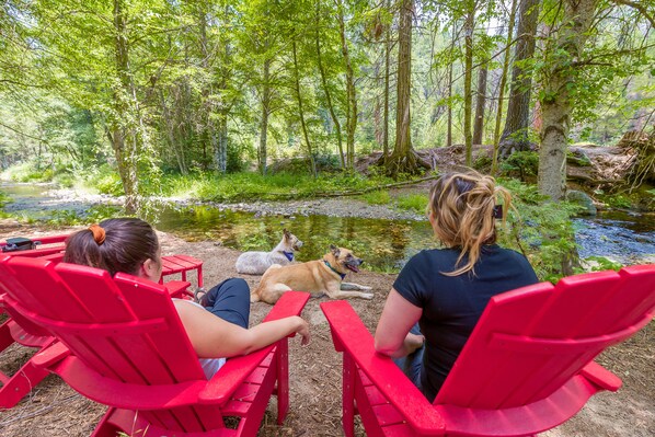 Three people seated on red Adirondack chairs facing a pond with two dogs nearby.