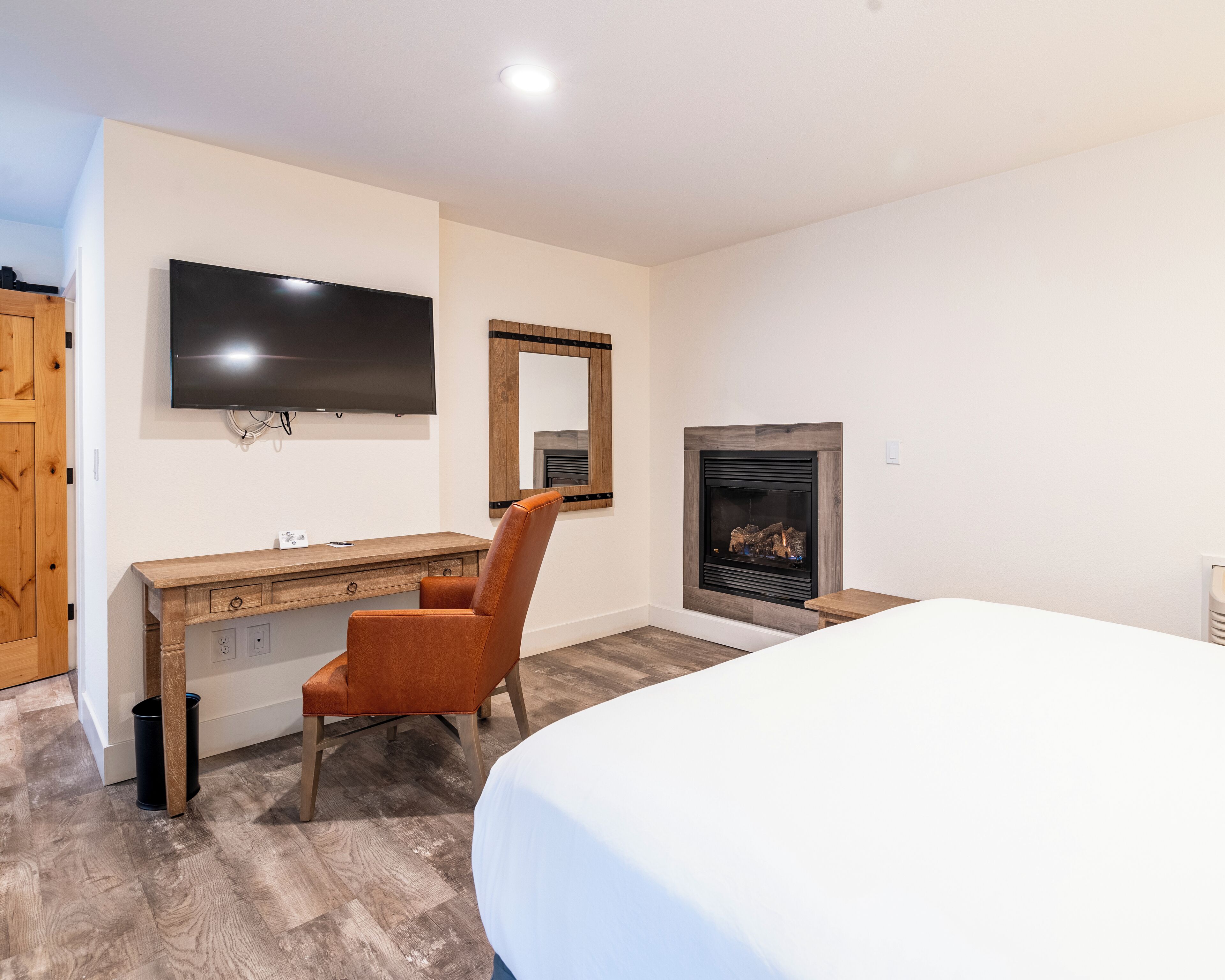 A hotel room with a wooden desk, a chair, a bed, a television, and a fireplace.