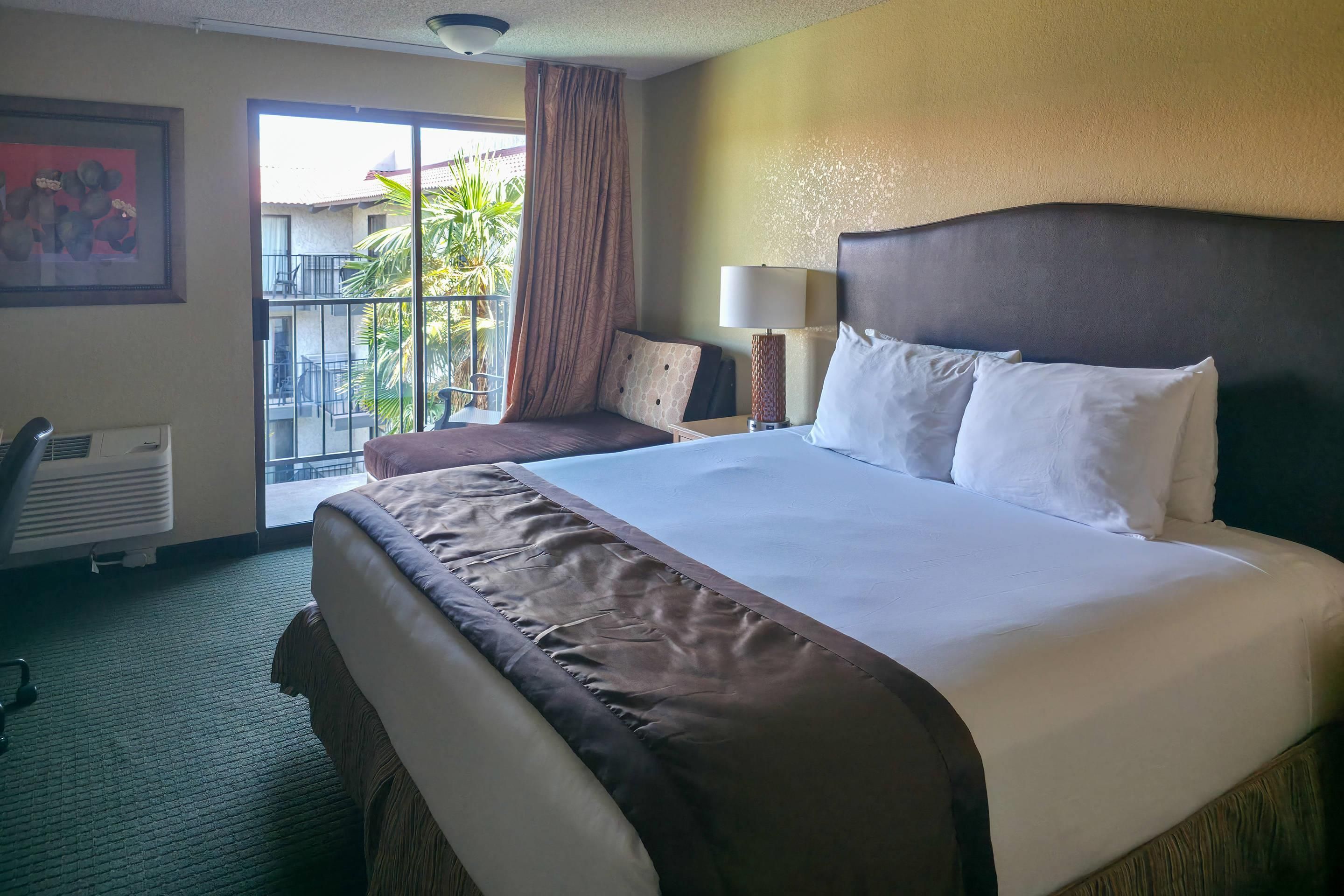 A hotel room with a large bed, a desk, a chair, a window with curtains, and a wall-mounted air conditioner.