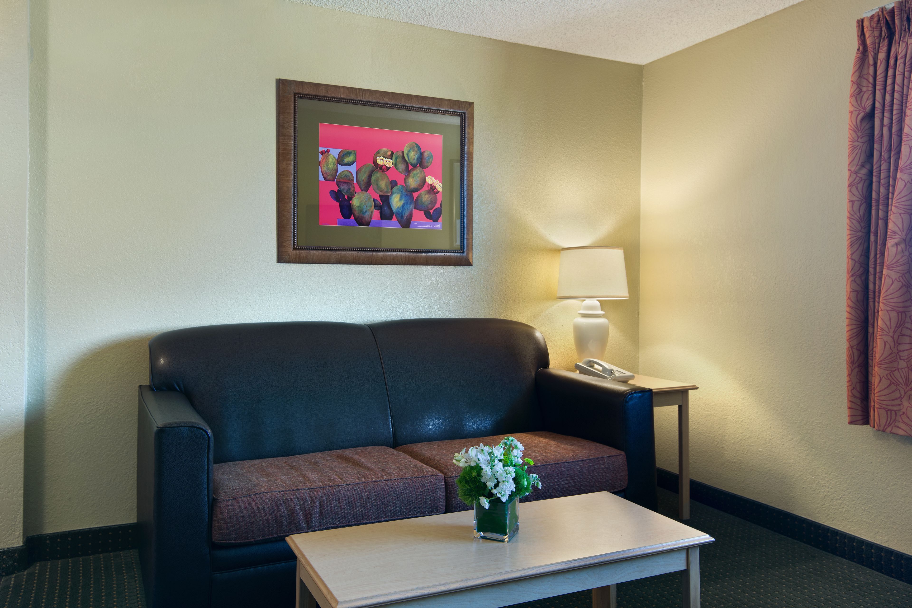 A hotel room with a black leather sofa, a small table with a vase of flowers, a framed picture on the wall, a lamp, and a telephone on a side table.
