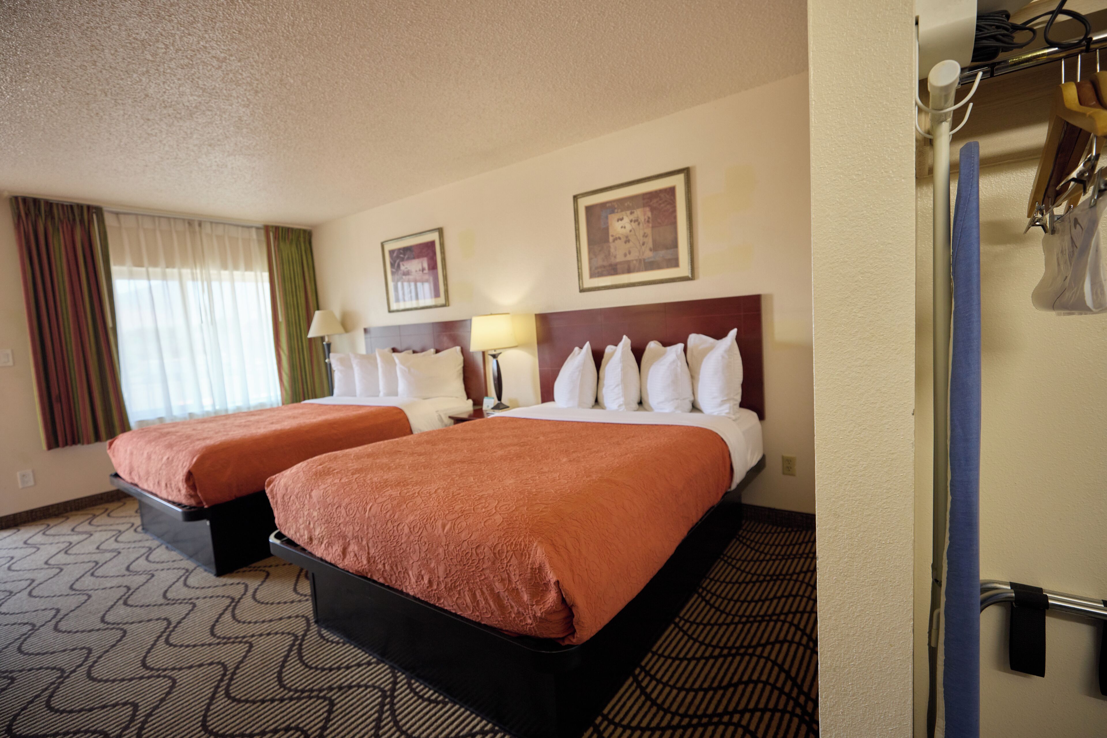 Quarto de hotel com duas camas, carpete estampado, cabeceira, janela com cortinas e luminária fixada na parede.