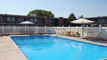 A swimming pool with lounge chairs and umbrellas, surrounded by a white picket fence, with a building in the background.