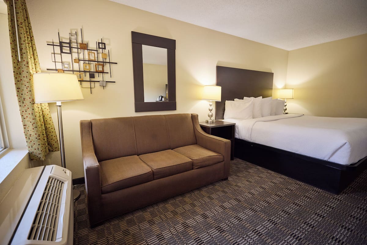 Une chambre d’hôtel avec un canapé marron, un lit avec du linge de lit blanc, un grand miroir et deux lampes de chevet.