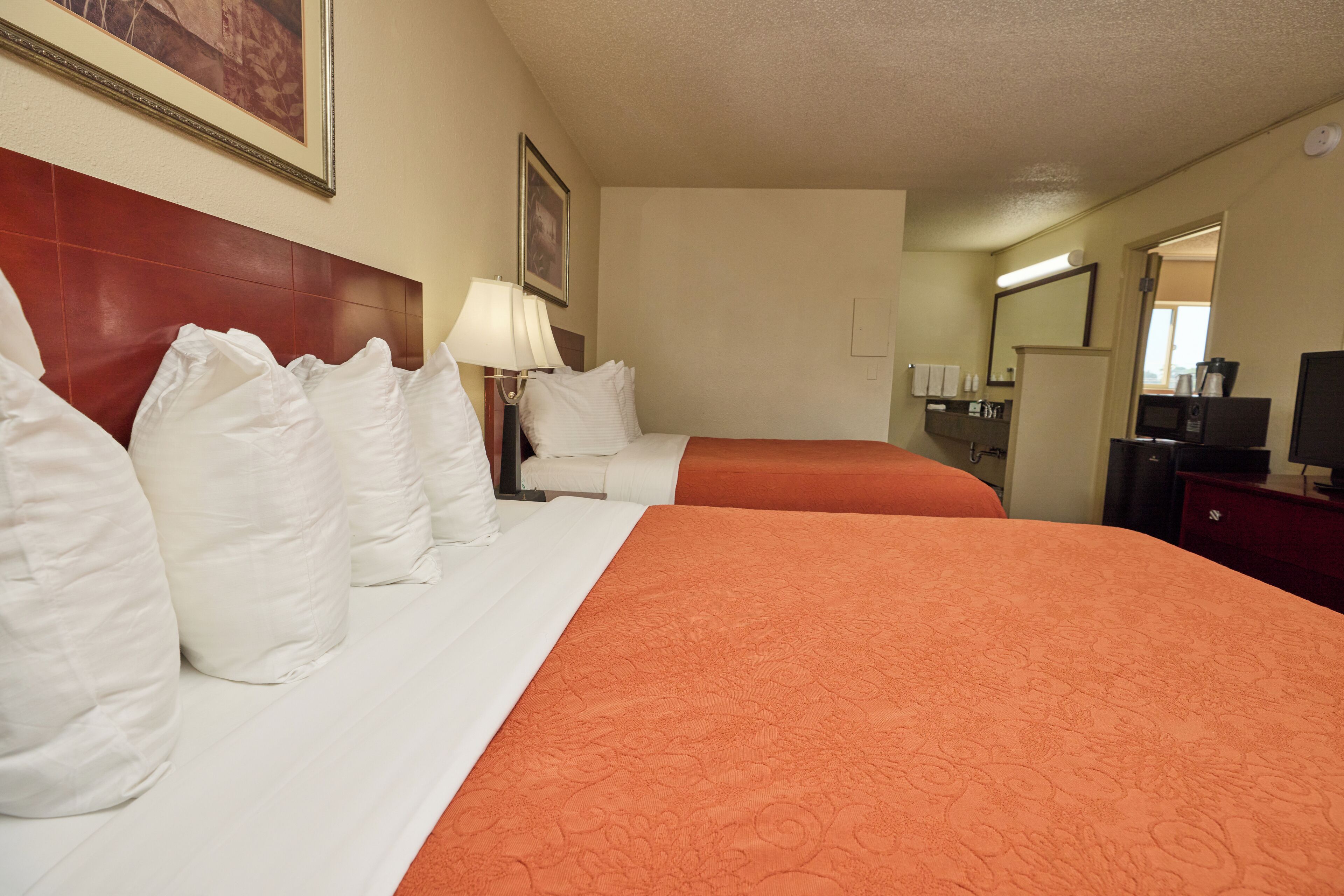 A hotel room with two beds, white bedding, an orange bedspread, a nightstand with a lamp, a dresser, a mirror, and a coffee machine.