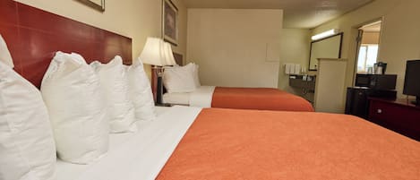 A hotel room with two beds, white bedding, an orange bedspread, a nightstand with a lamp, a dresser, a mirror, and a coffee machine.