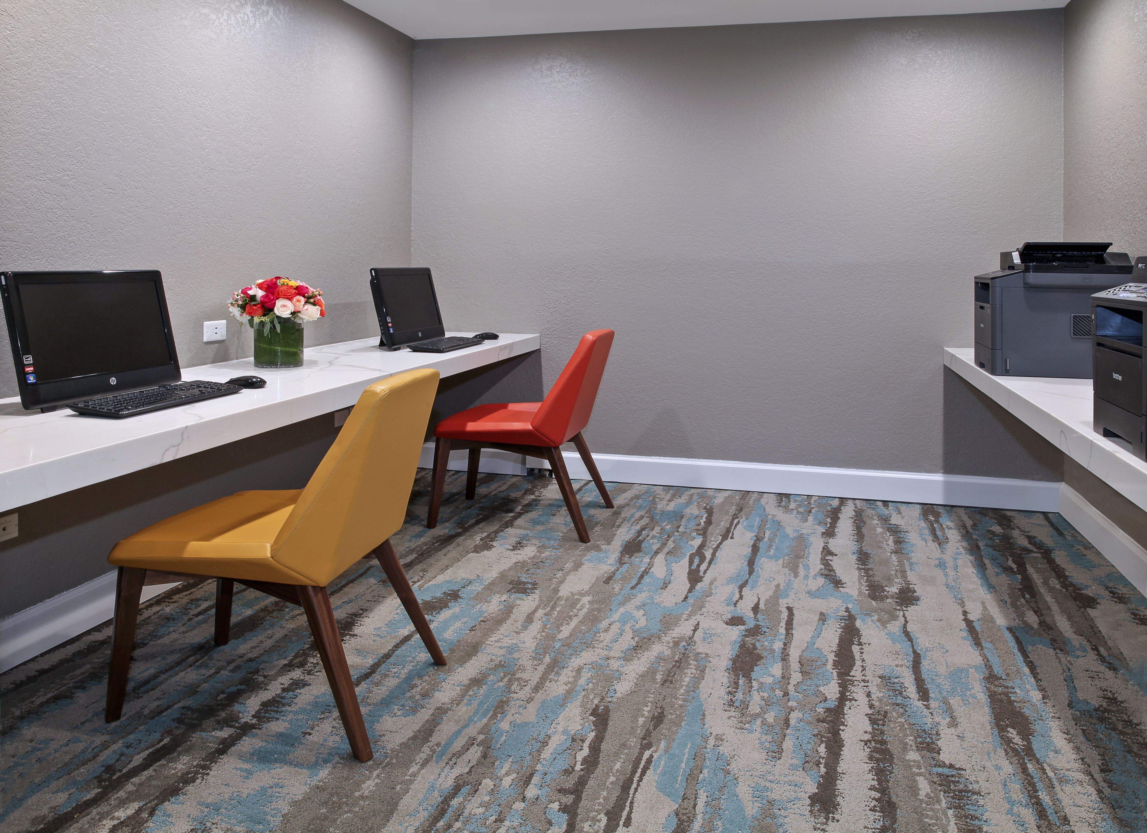 A hotel room with a desk, two chairs, a printer, and a vase with flowers.