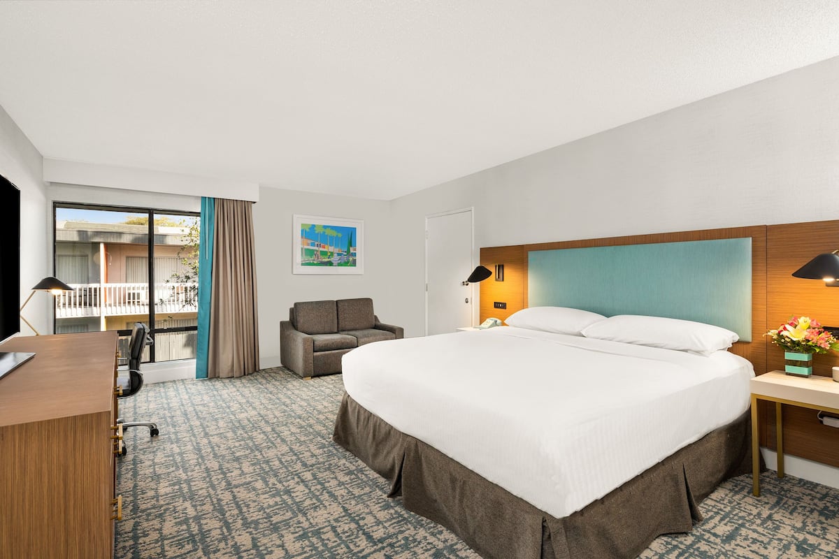 A hotel room with a large bed, a desk, a chair, a small table with a lamp, and a window with curtains.