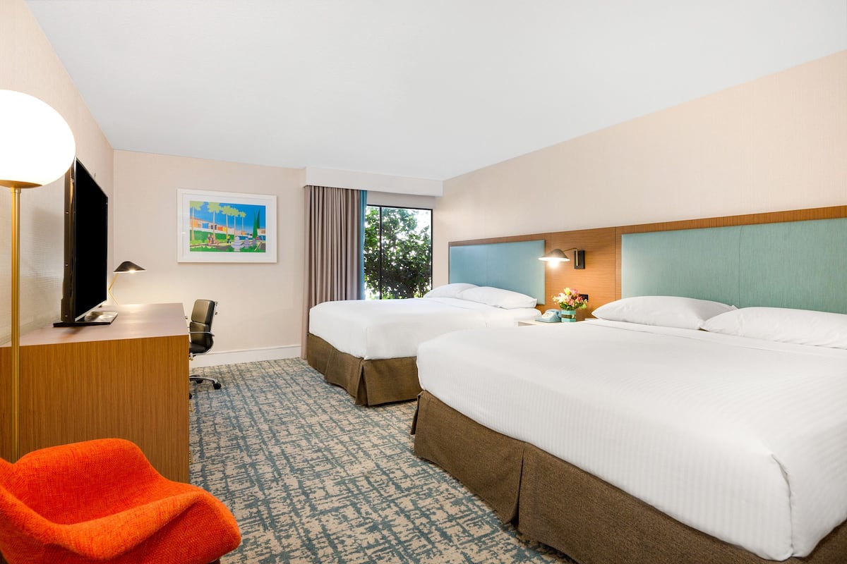 A hotel room with two beds, a desk, a chair, and a window with a view of trees.