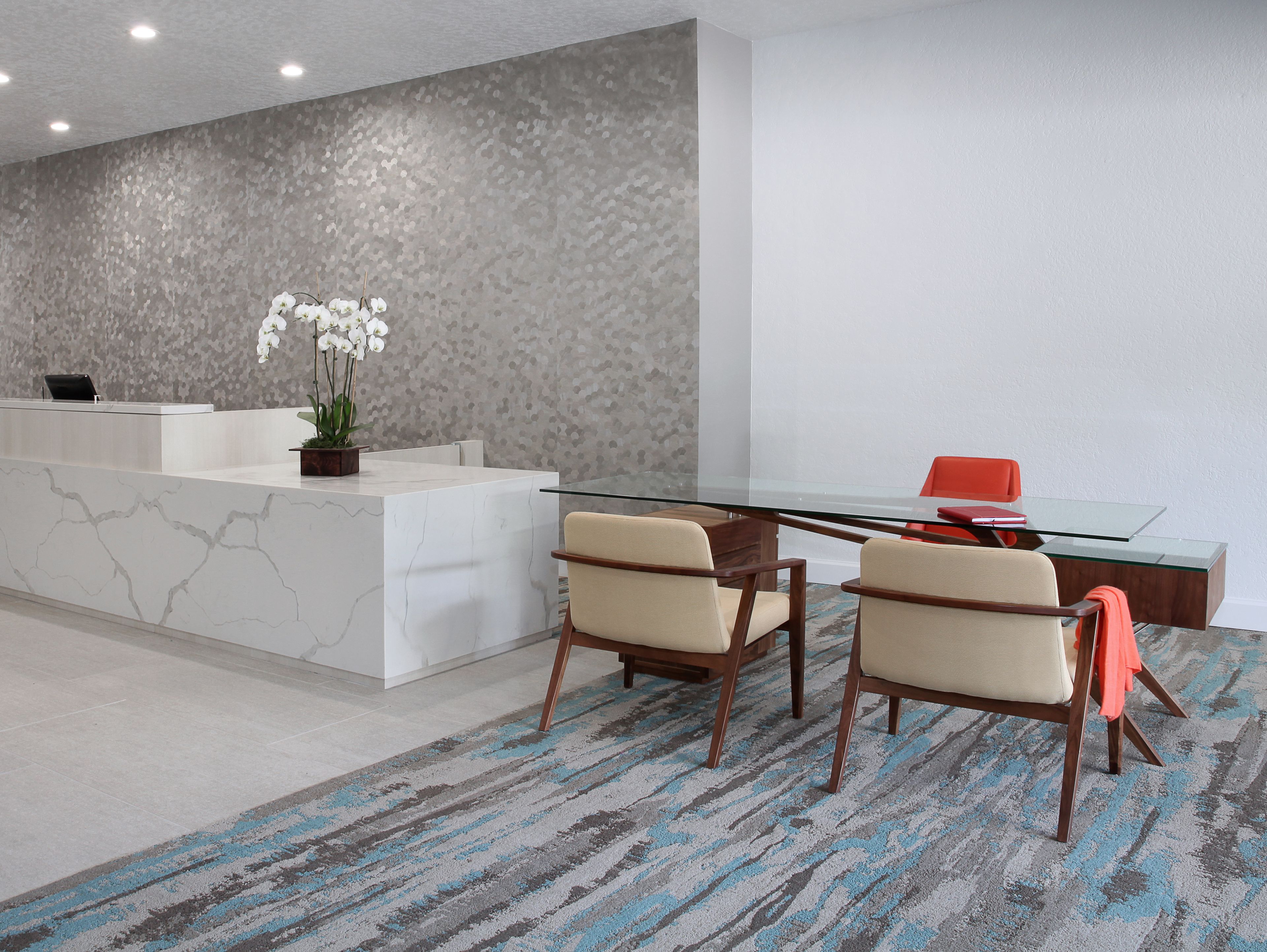 A reception area with a marble counter, a potted orchid, two beige chairs, and a glass table.